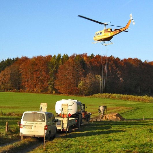 Hubschrauber und Hilfs-Fahrzeuge vor einem Waldstück. Der Hubschrauber wird mit Kalk beladen.