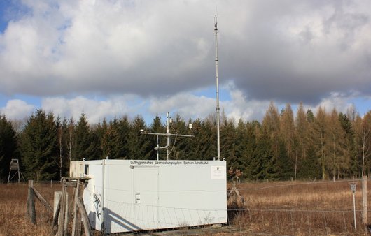 weißer Container mit Messinstrumenten außen auf einer Fläche vor einem Wald