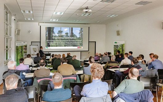 Zuhörende sitzen im Saal und schauen auf eine Leinwand mit Vortragsfolien.
